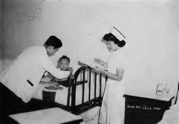 An evacuee doctor examining a baby while an evacuee nurse writes the medical record